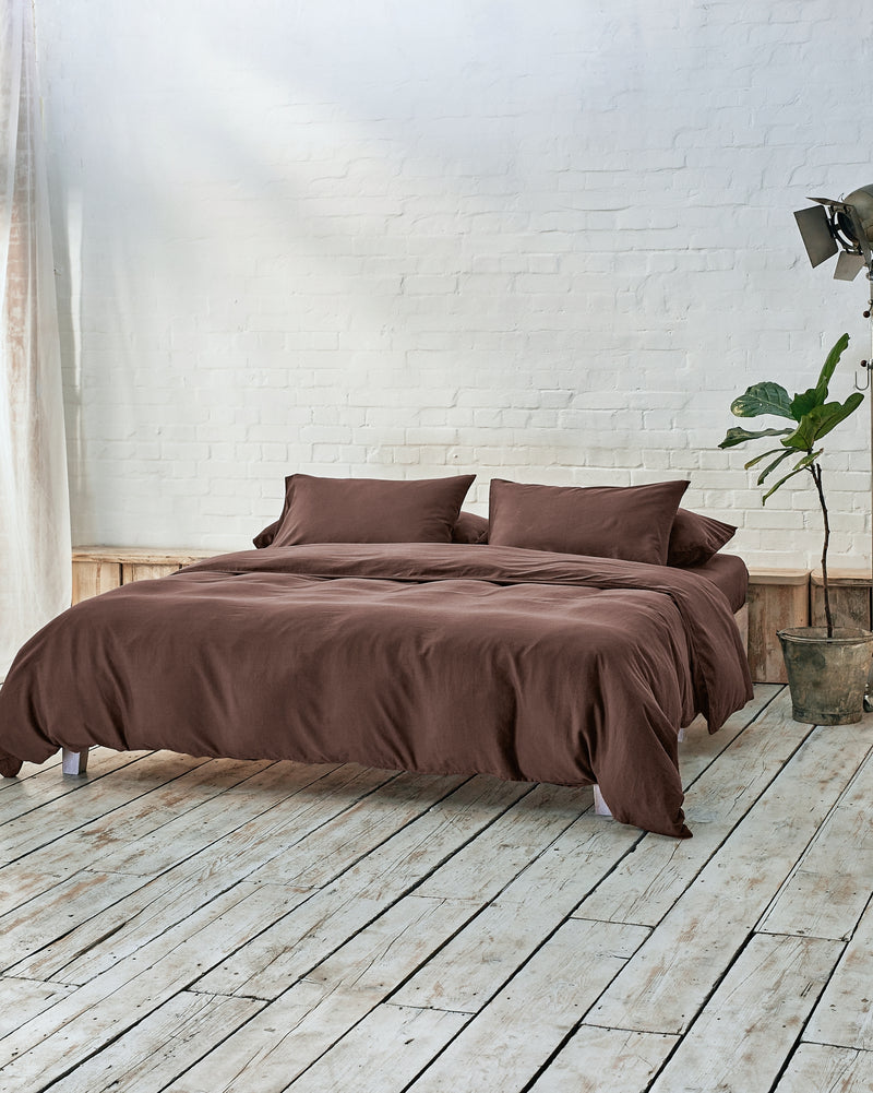 lifestyle image of acorn brown bedding set, situated within a modern bedroom with rustic wood flooring and white exposed brick walls.