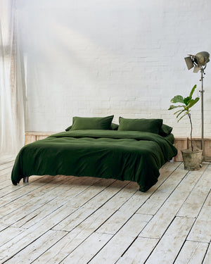 side shot of dark green bedding set in a modern bedroom with rustic wooden flooring, white brick walls, a tall plant and vintage lamp. 