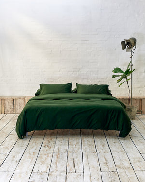lifestyle image of dark green bedding, set in a modern bedroom with rustic wooden flooring and white brick walls. 