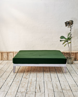 dark green flat sheet, lifestyle shot set in a modern bedroom with rustic wood flooring and white brick walls. 
