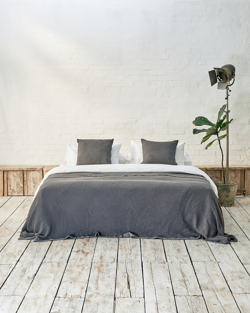 front facing lifestyle image of dark grey waffle throw and matching scatter cushions on a cloud grey bed. 